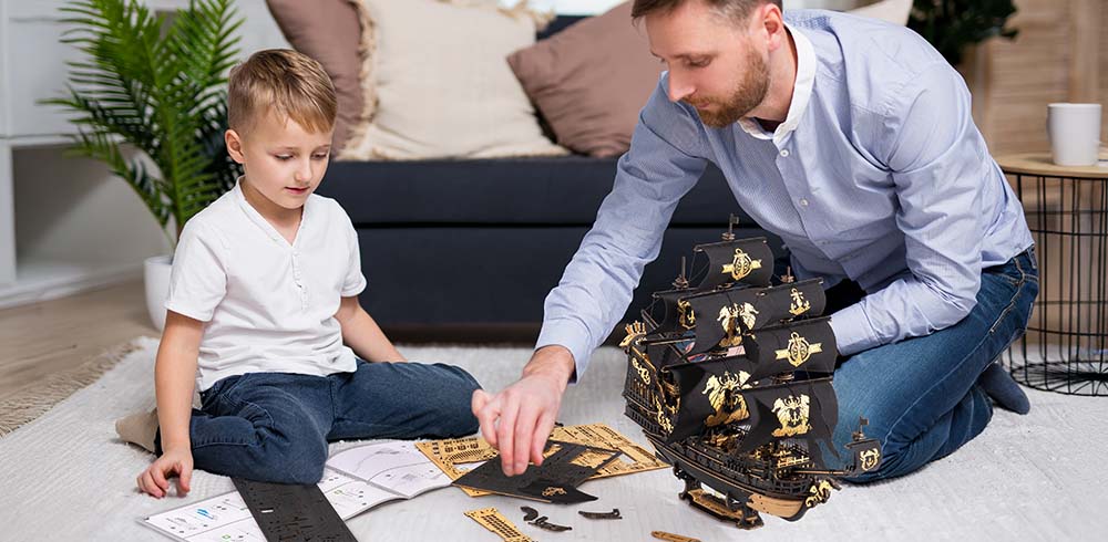 A father and young boy assemble the black and gold ROKR Seahorse Barque wooden pirate ship on a white living room rug with building pieces spread around