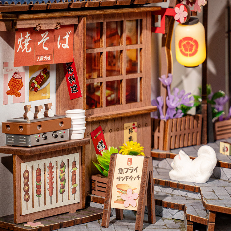 Close-up of Rolife Fukuro-cho book nook yakisoba stall at dusk, showing food menu signage, a glowing lantern, a white cat figurine, and cobblestone path.