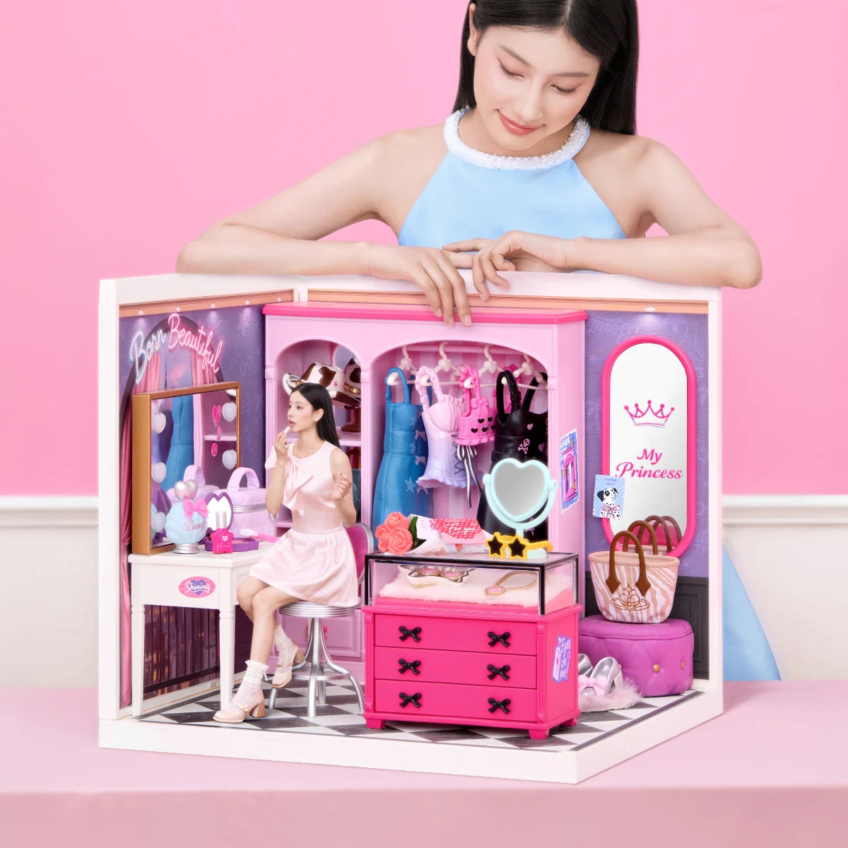 Miniature dressing room with vanity, clothing rack and pink drawers, with a person leaning over it against a pink background.