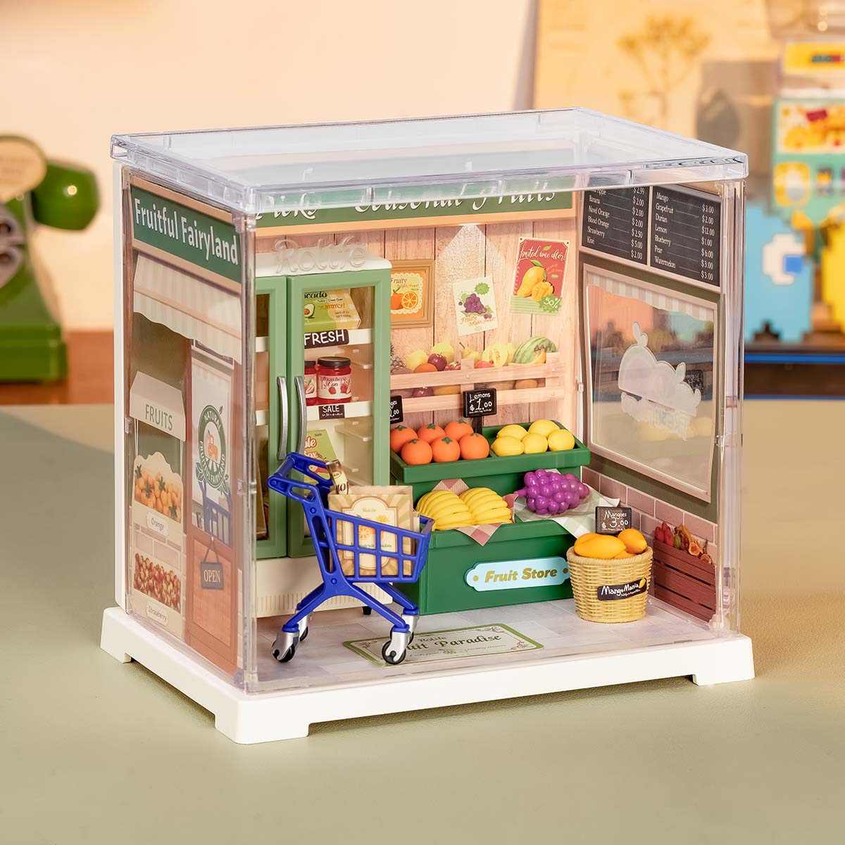 Rolife Nature Energy Fruits DWS01B lifestyle view showing the green fruit store miniature with pushable shopping cart inside its clear acrylic display case on a desk.
