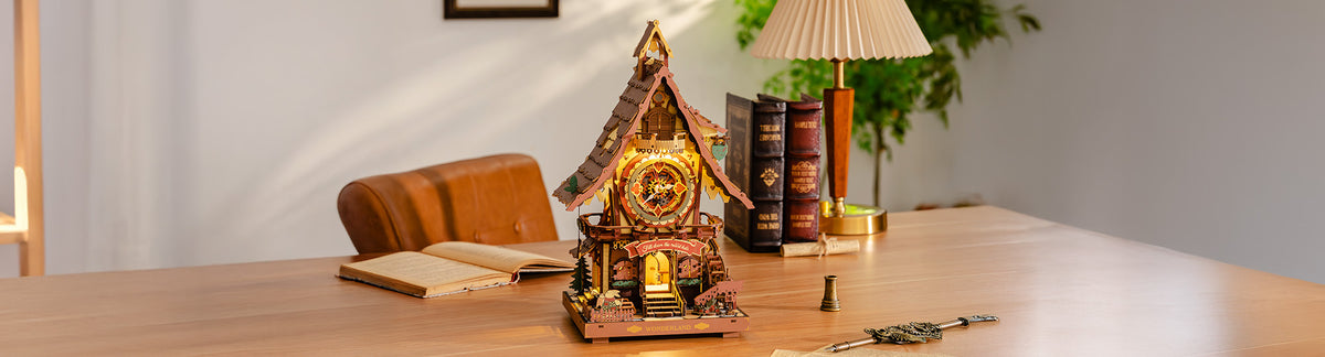Robotime Online Wonderland themed banner showing a glowing ROKR wooden cuckoo clock house with intricate gear and floral motifs beside stacked books and a table lamp on a wooden desk.