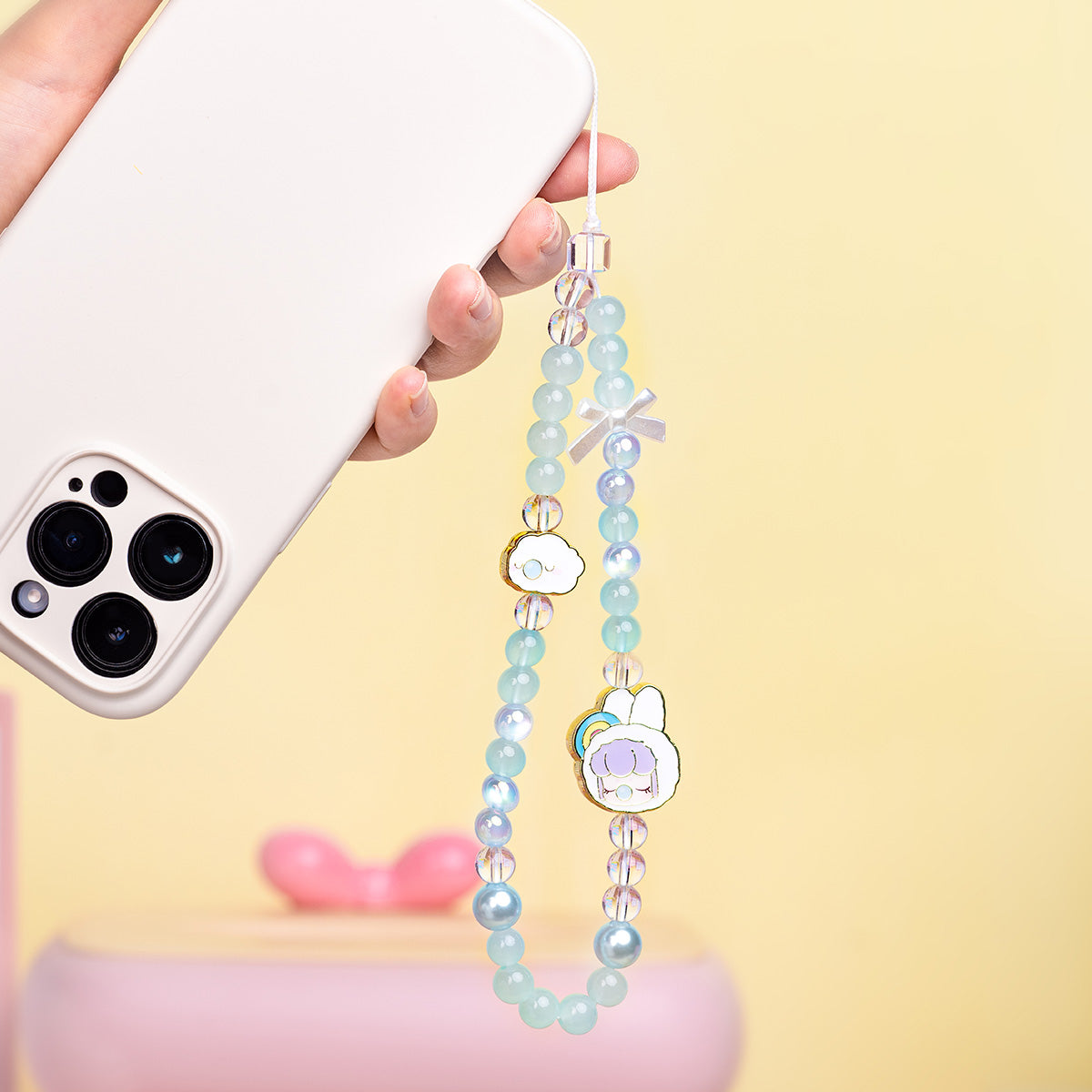 Hand holding a phone with a Rolife Nanci Fluffy Cloud mint bead lanyard hanging down, showing cloud and bunny enamel charms against a yellow background.