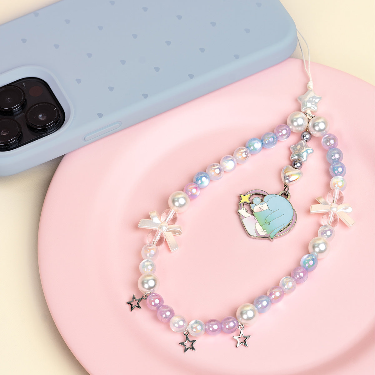 Rolife Nanci Sweet Time Hidden Honey Air lanyard coiled on a pink plate beside a blue phone case, showing iridescent pearl beads and illustrated enamel charm.