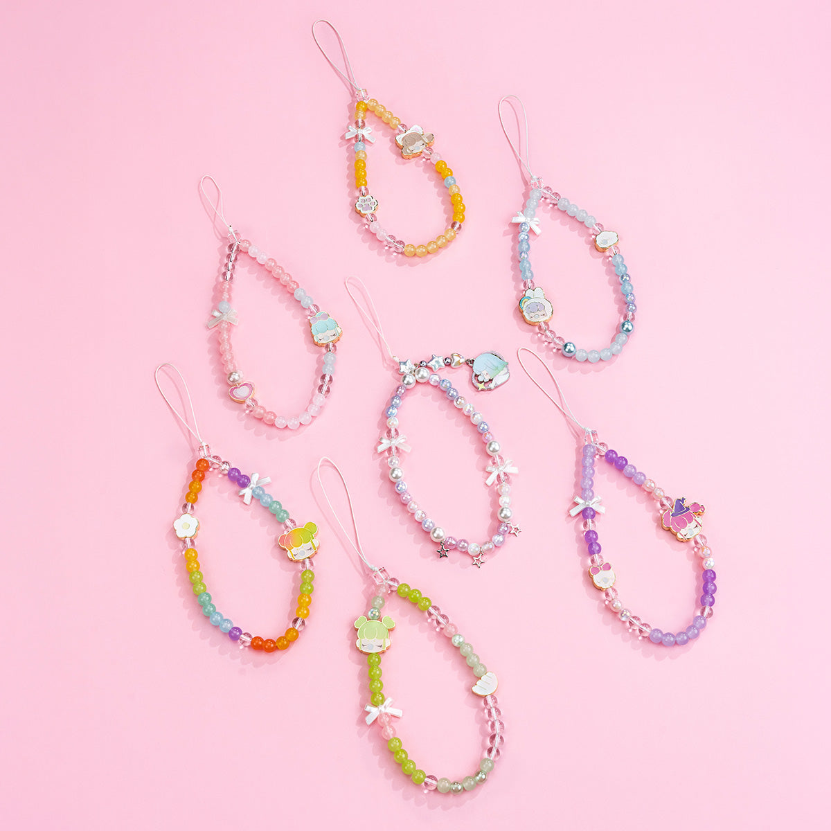 Seven Rolife Nanci Sweet Time beaded phone lanyards in rainbow, blue, pink, green, purple and pearl colors laid out on a pink surface.