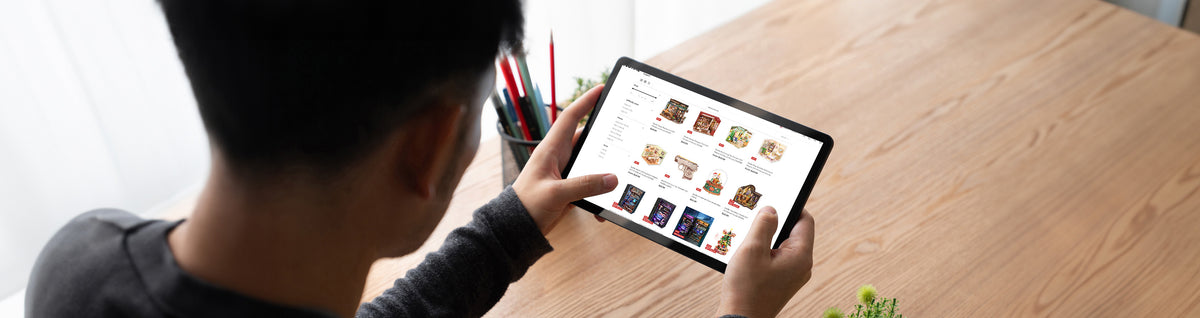 A person browsing the Robotime online store catalog on a tablet at a wide wooden desk, showing a grid of Rolife and ROKR product listings with prices and a small succulent plant nearby.