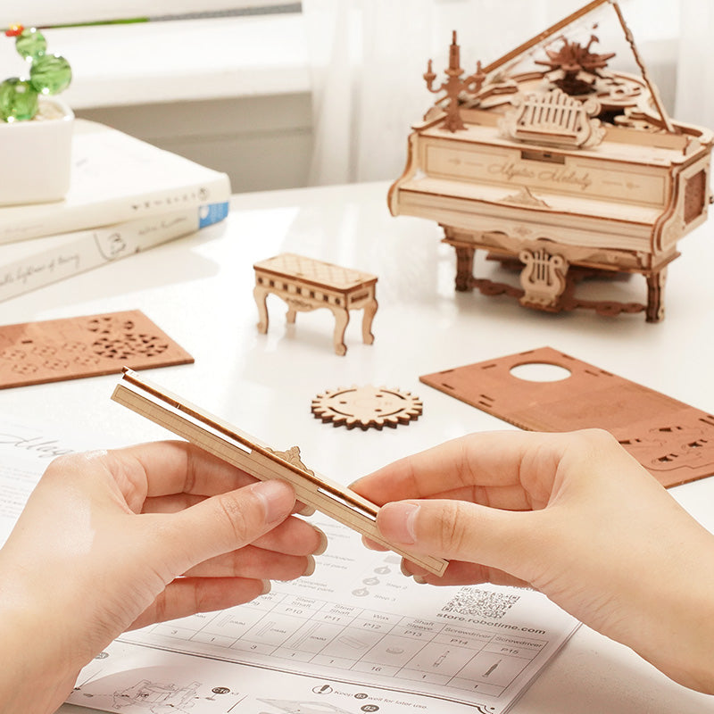 Hands assembling the ROKR Mystic Melody grand piano music box with wooden laser-cut parts and instruction sheet spread on a desk beside the finished piano model