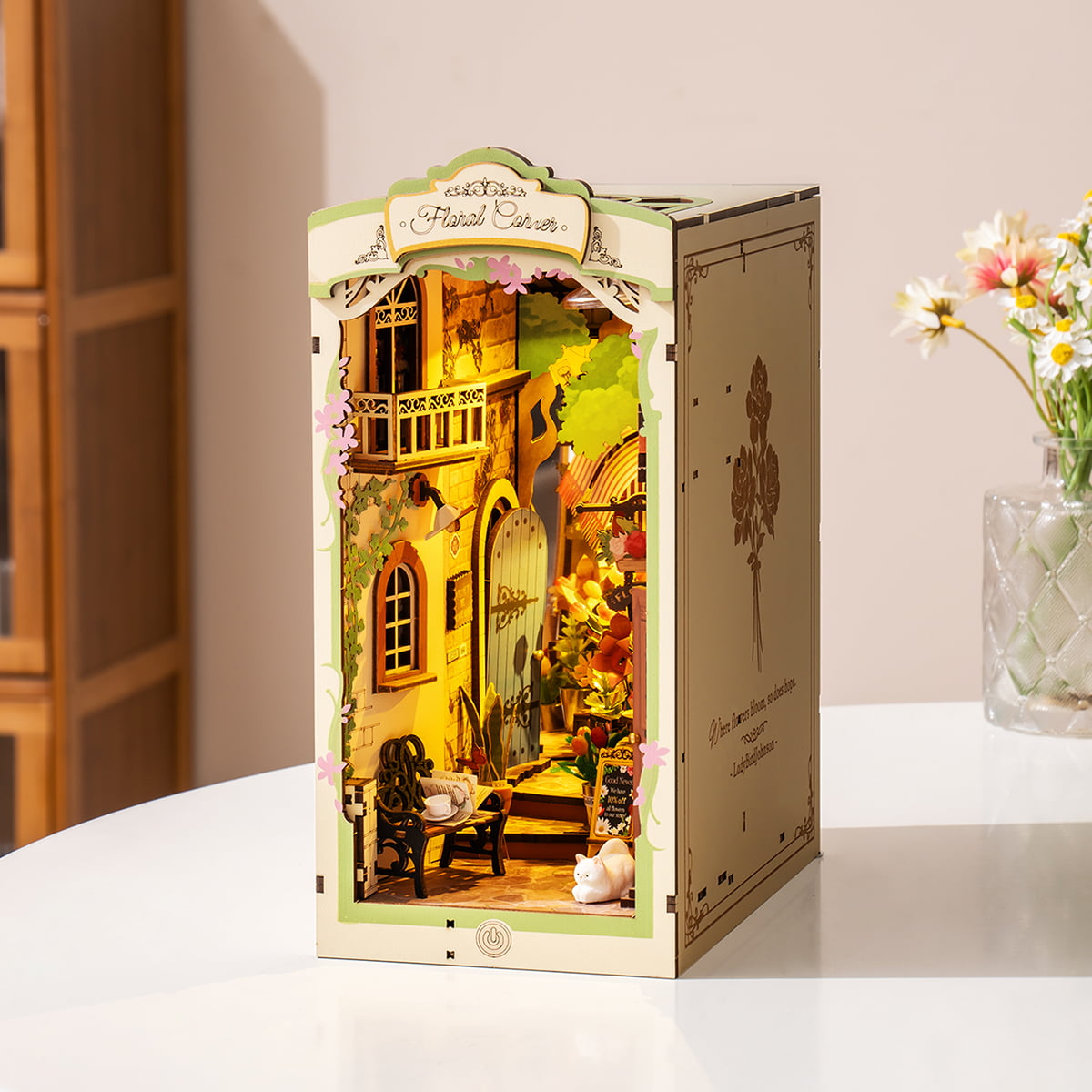Rolife Floral Corner TGB09 Book Nook standing on white table beside real flowers showing the full illuminated miniature flower shop interior