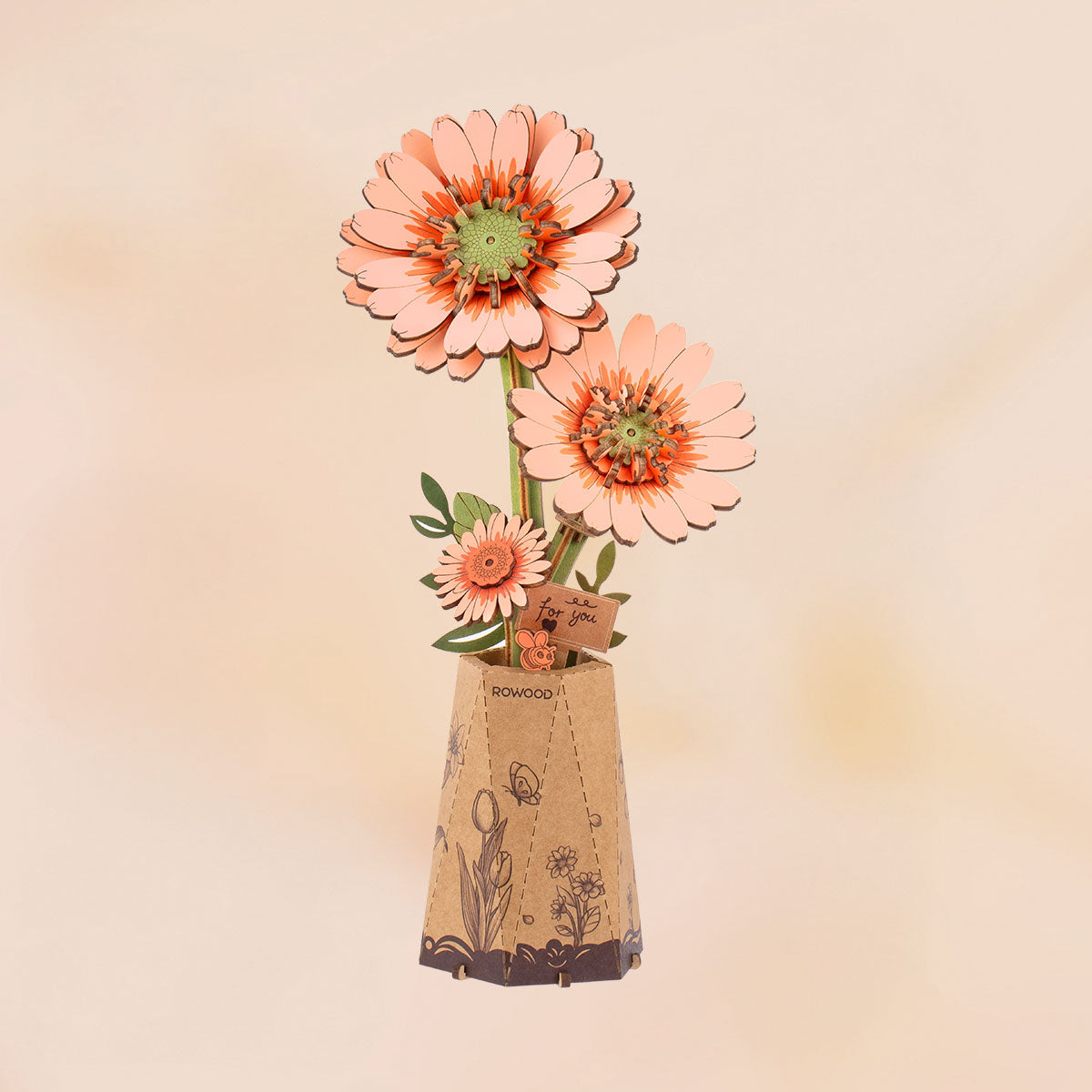 Rowood DIY wooden flower bouquet TW111 completed gerbera daisy model showing two large peach and coral blooms with a smaller bud in a kraft paper pot on a peach background.