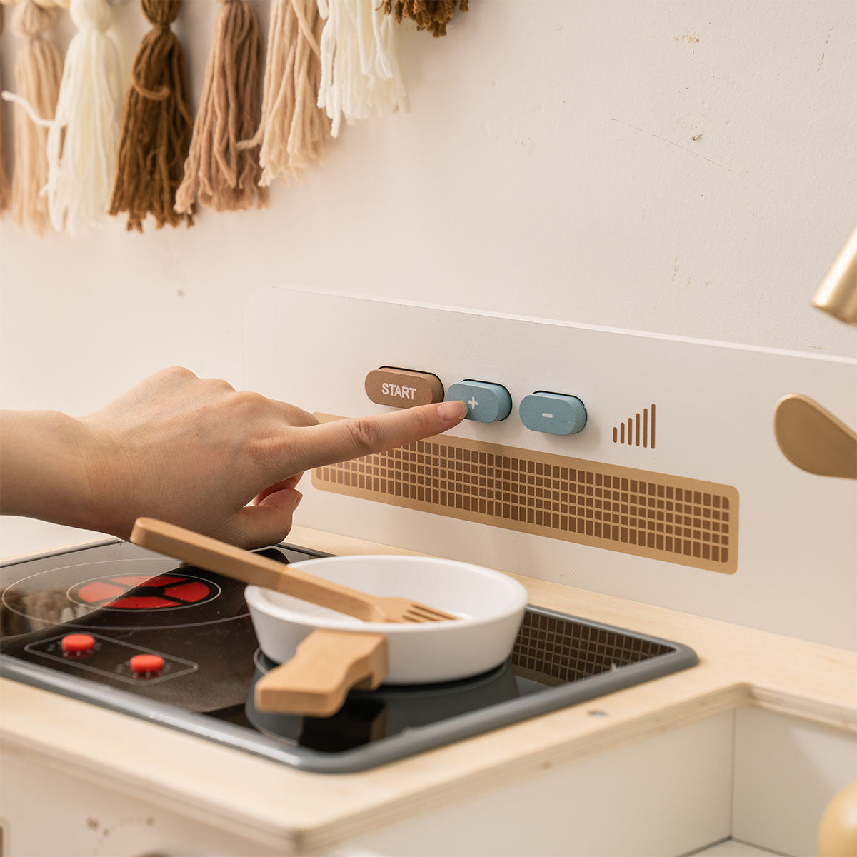 Child's hand pressing controls on ROBUD white wooden play kitchen stovetop with pan and wooden spatula.