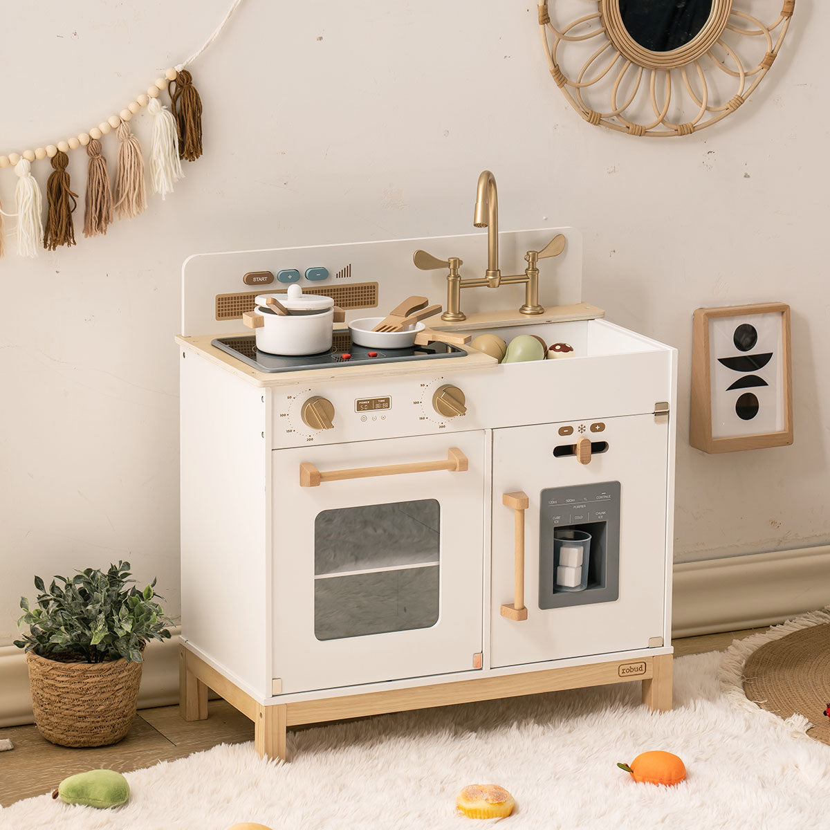 ROBUD white Montessori play kitchen angled view with macrame decor and autumn leaves on floor.