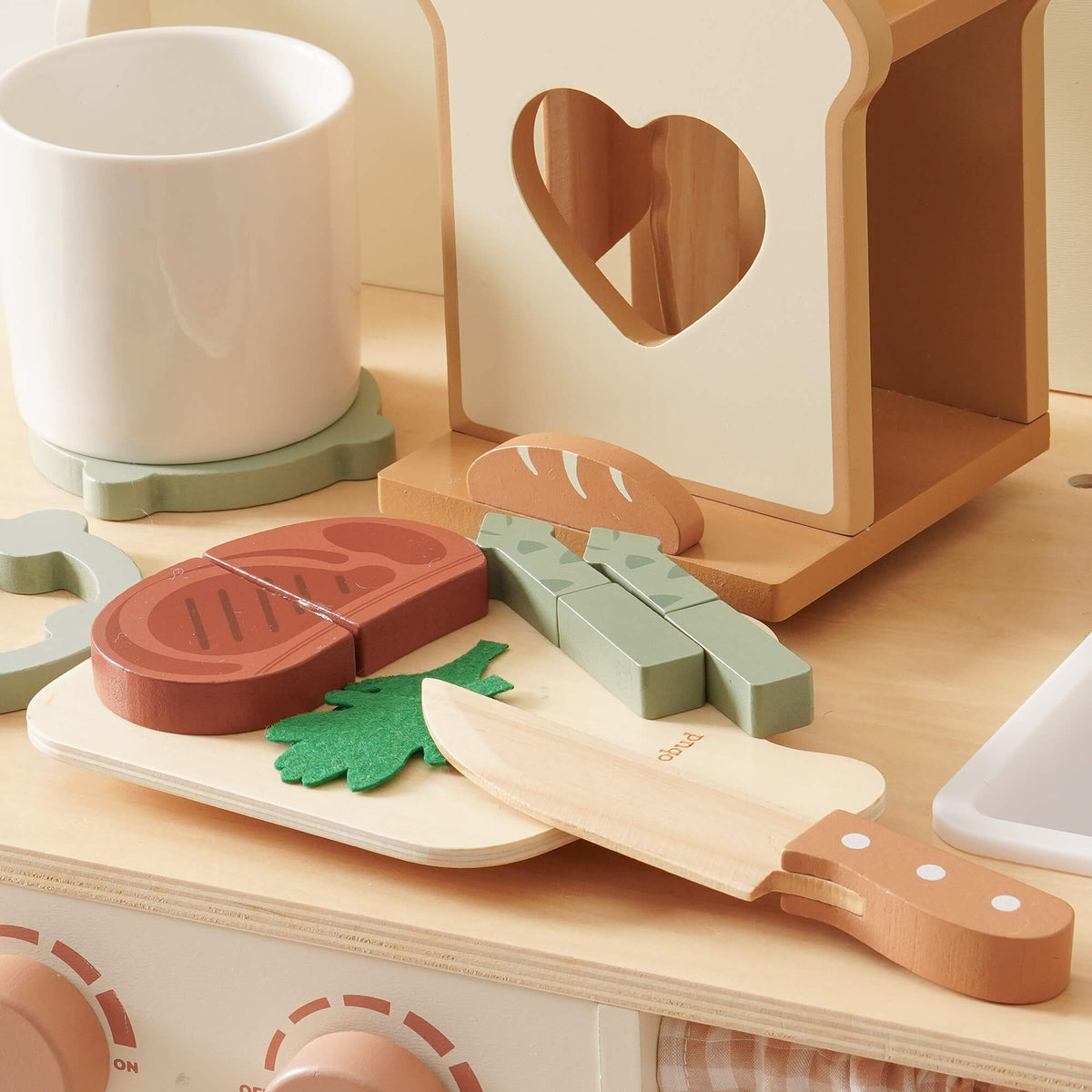 ROBUD wooden kitchen toy set close-up of natural birch cutting board holding dark red toy steak, green felt parsley, asparagus pieces and a sage-handled toy knife