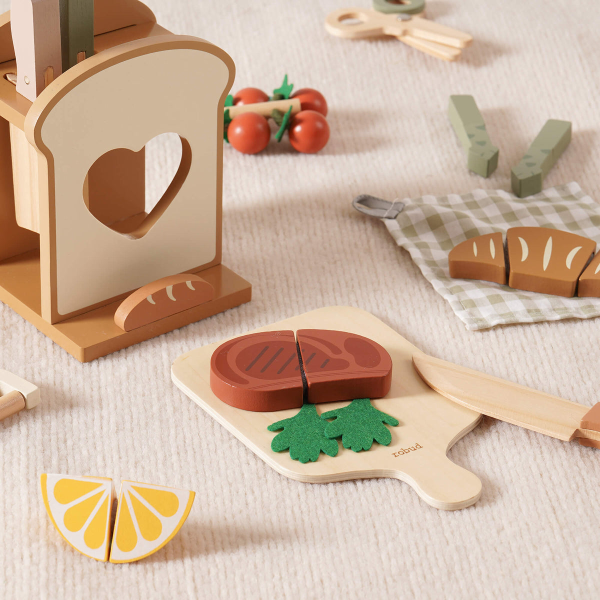 ROBUD wooden play kitchen scene with a cutting board holding toy steak and green felt parsley, a heart-shaped toast holder and scattered food accessories on cream carpet