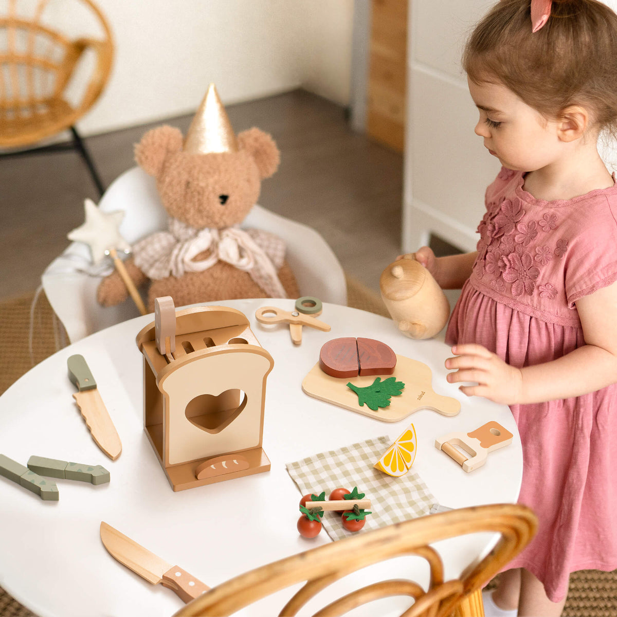 Young girl in pink dress playing with ROBUD wooden Mocha Cookware Set at a round table beside a teddy bear wearing a party hat, with toy steak and vegetables arranged around her