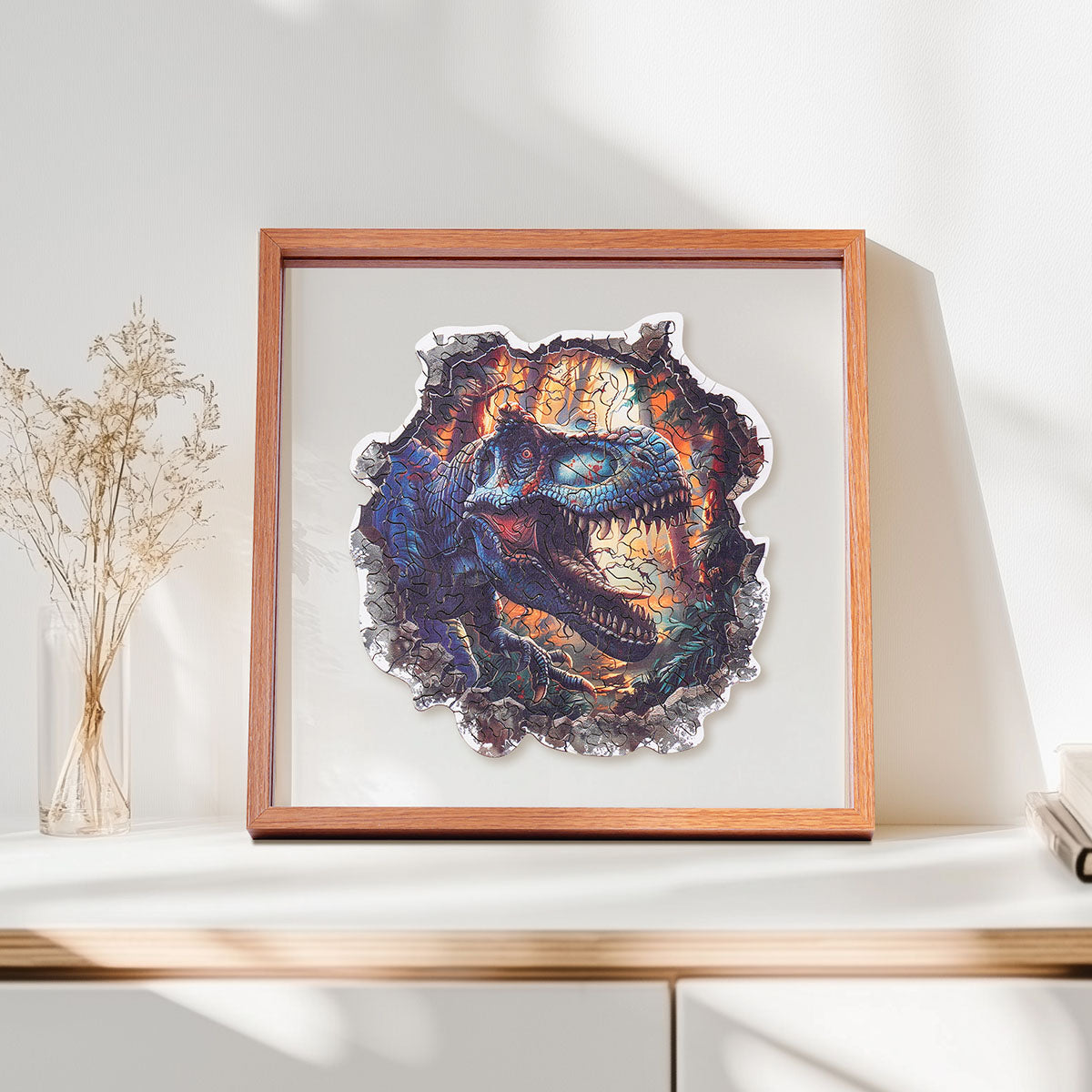 Rowood Wall Breaking T-Rex wooden jigsaw puzzle in a shadow box frame resting on a shelf beside dried flowers and books