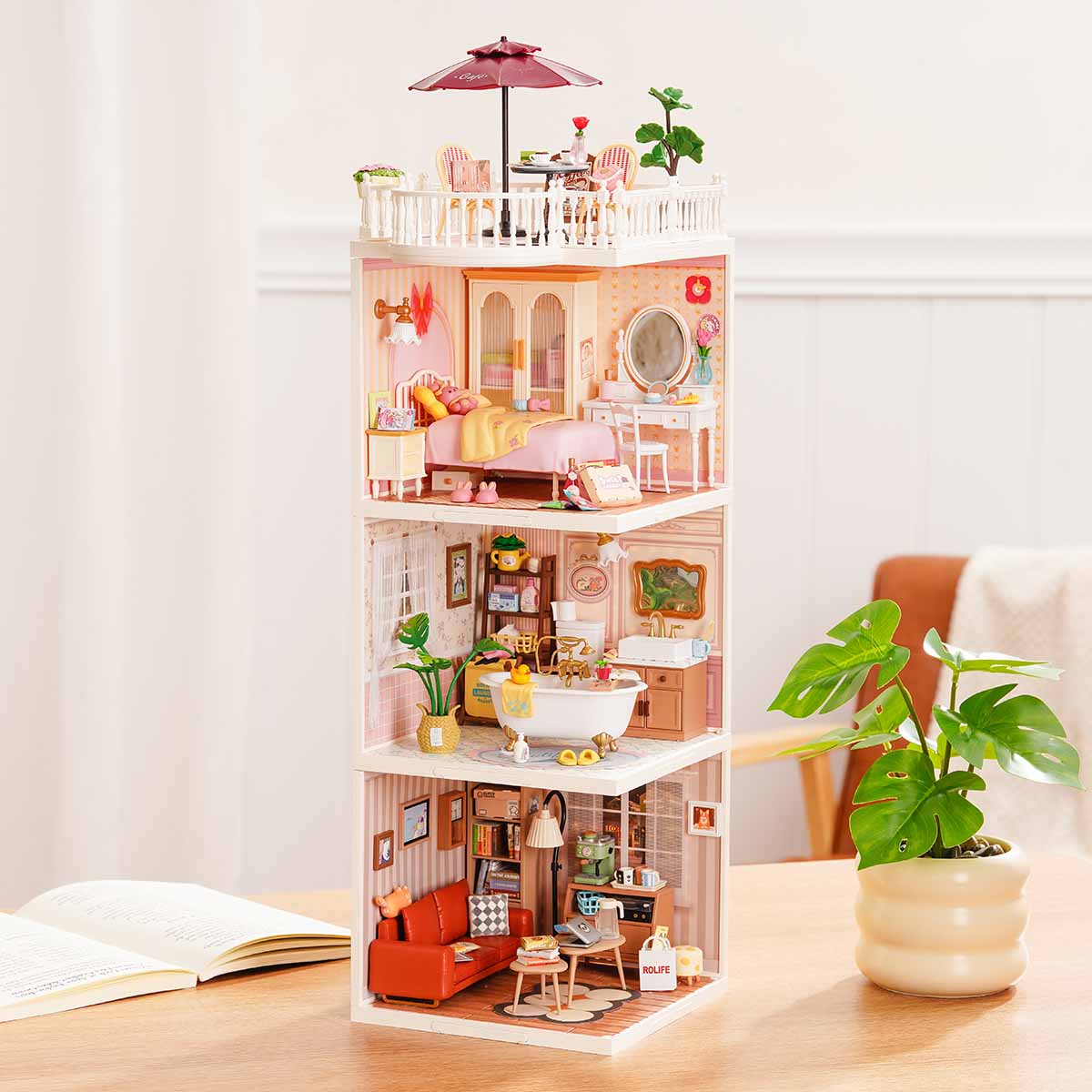 Rolife Super Creator Series 3-in-1 I Shape miniature tower showing three home rooms including pink bedroom, bathroom, and book-filled living room stacked with a glass roof on a table.