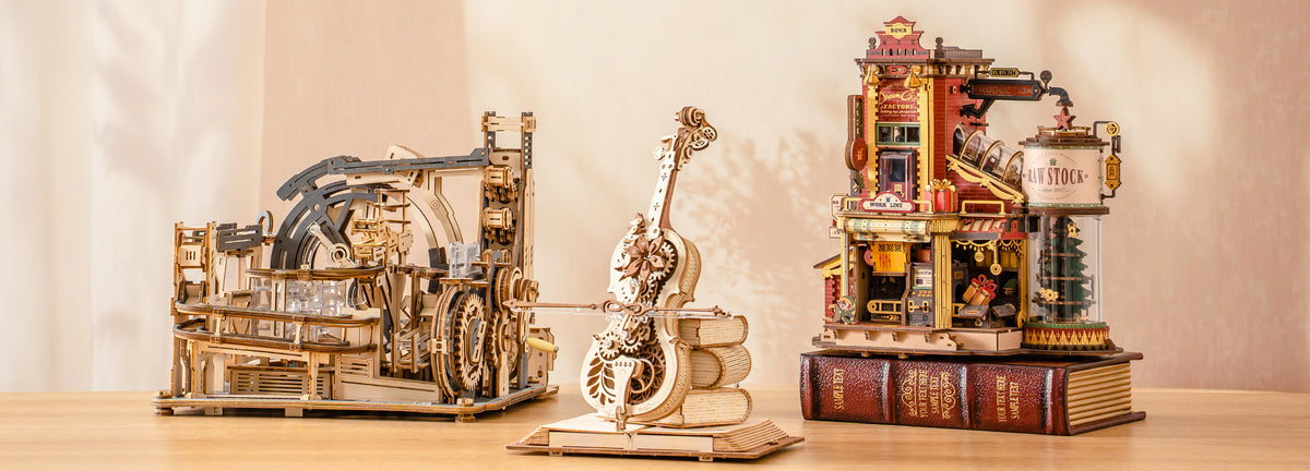 ROKR collection desktop banner showing three assembled mechanical wooden puzzle models including marble spaceport, dream gift factory, and cello music box on a warm beige shelf