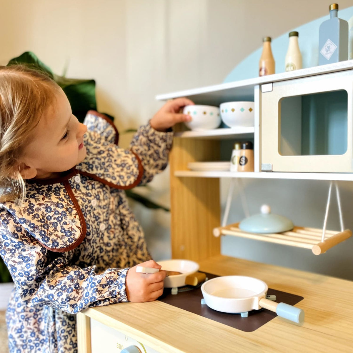 Toddler in a floral dress reaching up to the shelf of the ROBUD wooden kitchen play set WCF14 with toy bowl and bottles visible on the upper shelves and a pan on the cooktop