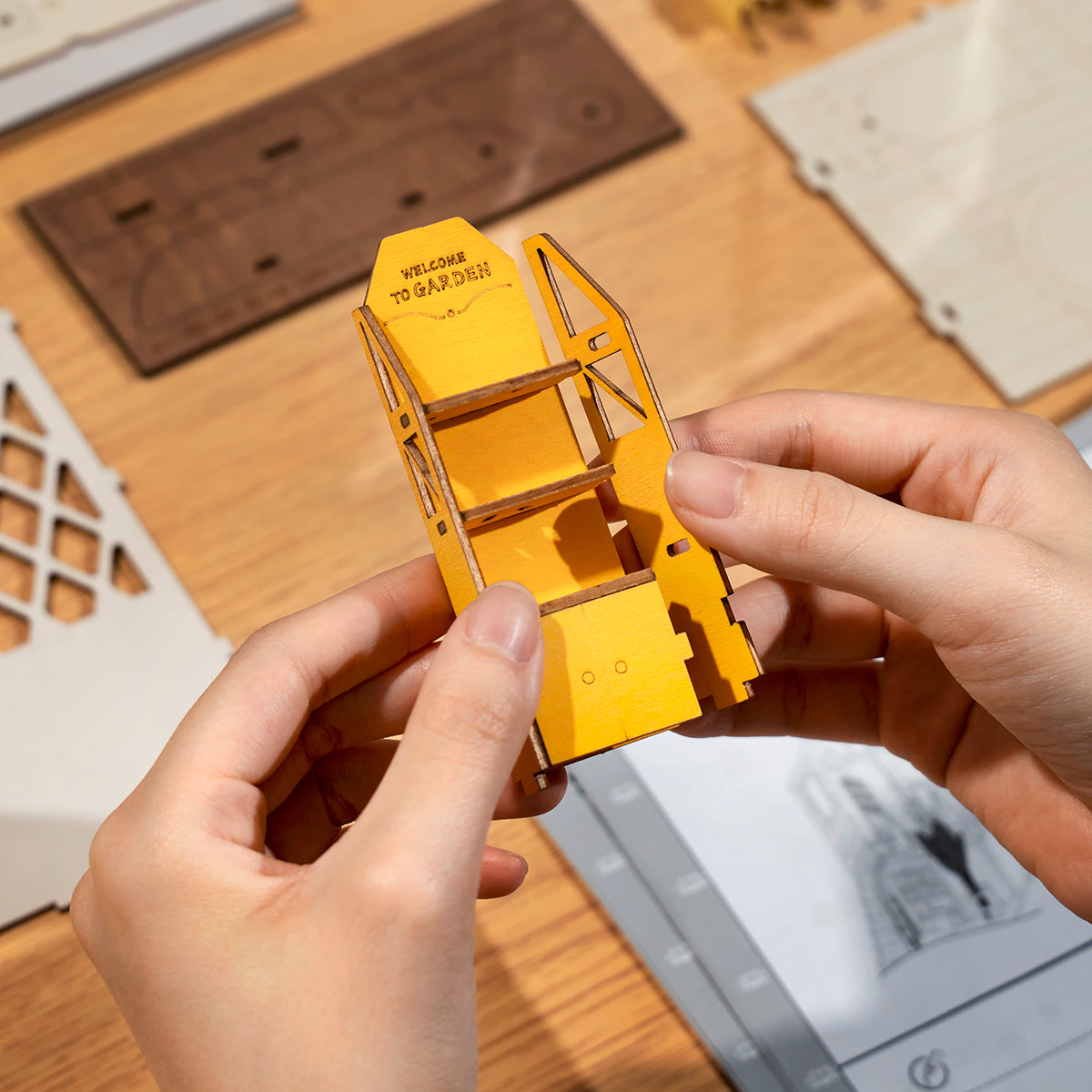 Hands assembling a small yellow Rolife miniature wooden structure piece with paper instructions nearby