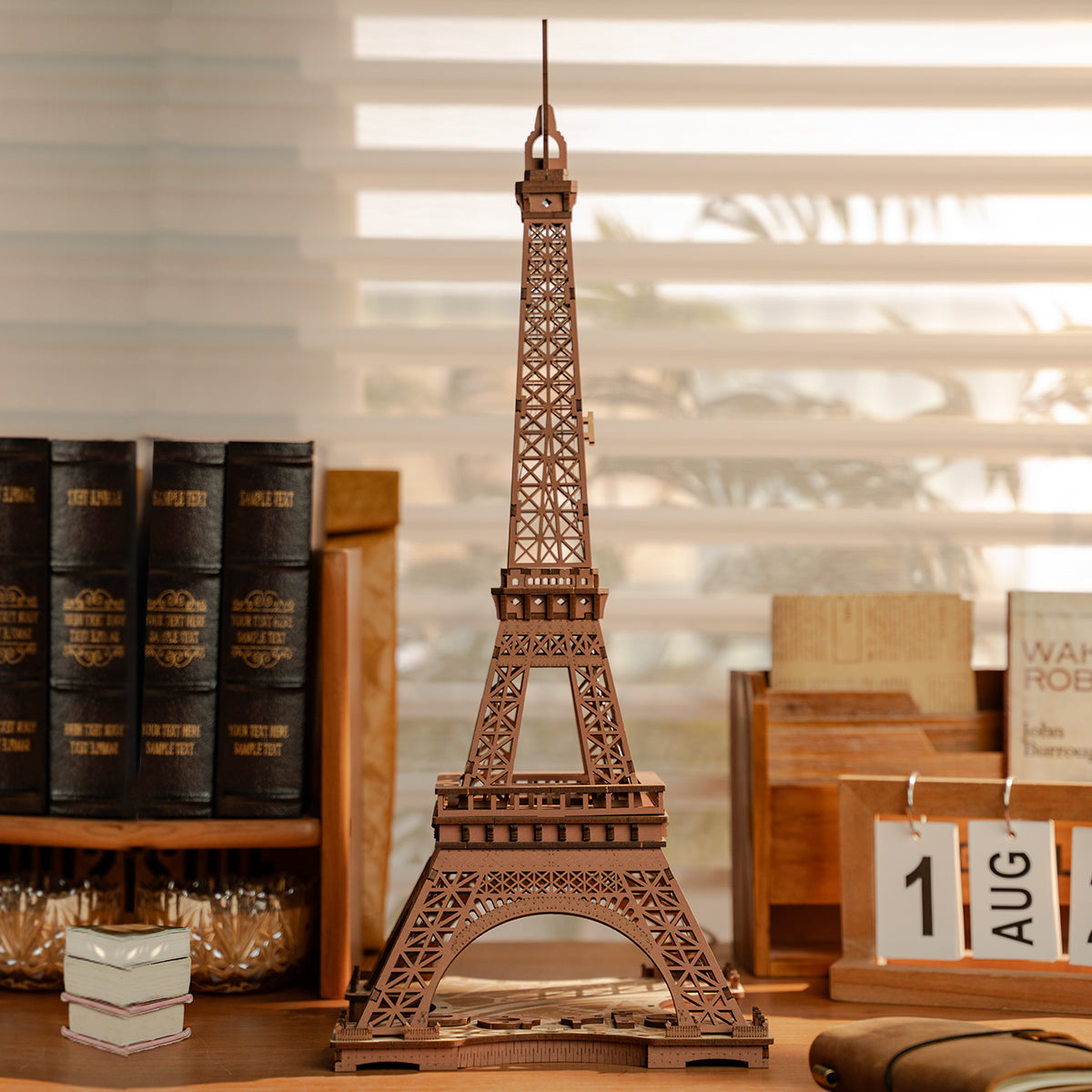 Completed Rolife Eiffel Tower wooden model in dark walnut tone displayed on a shelf beside leather books and an August desk calendar