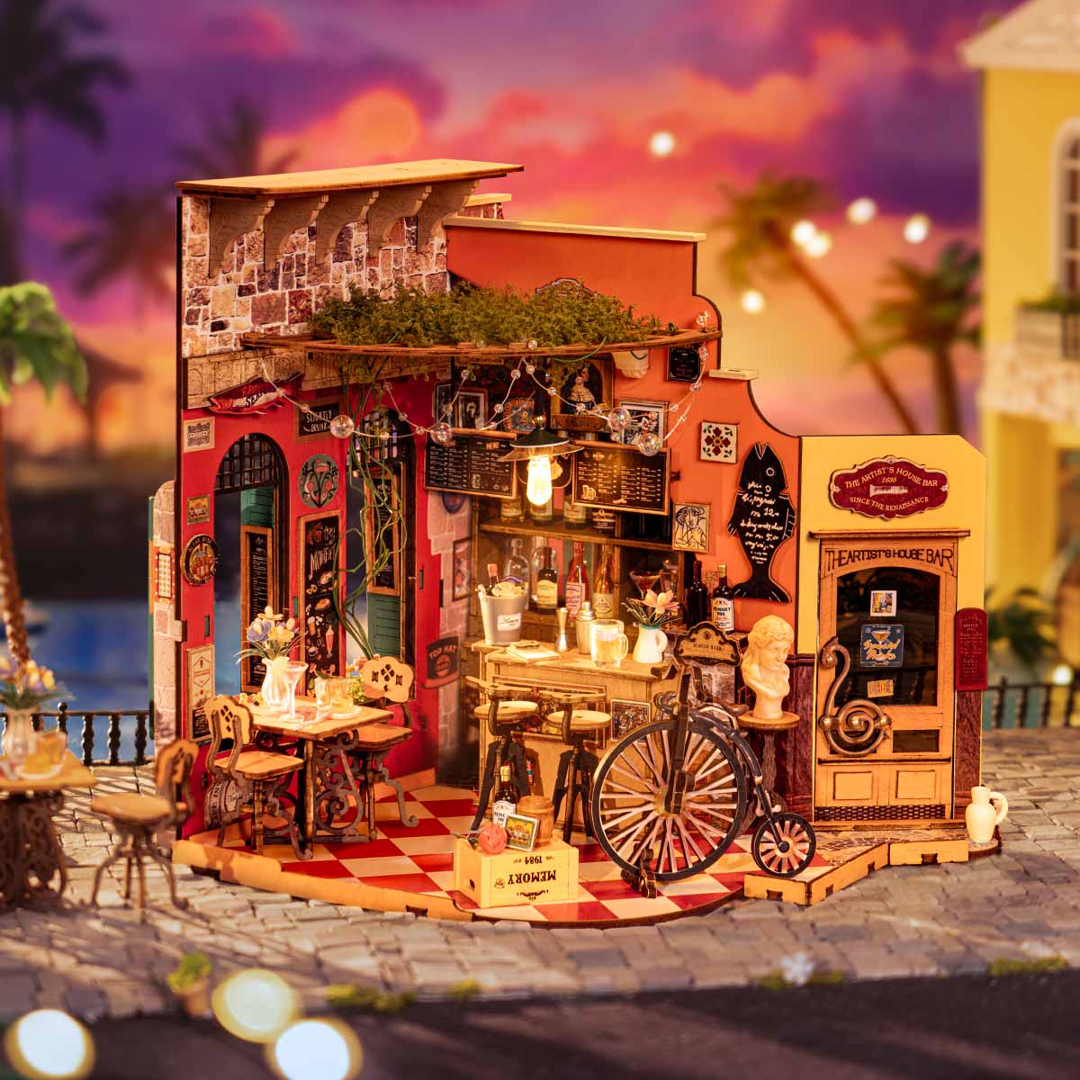 Rolife Cheers Bar and Dining DIY Miniature House DG173 fully assembled on a cobblestone plaza at sunset with warm string lights glowing over the bar counter and penny-farthing bicycle