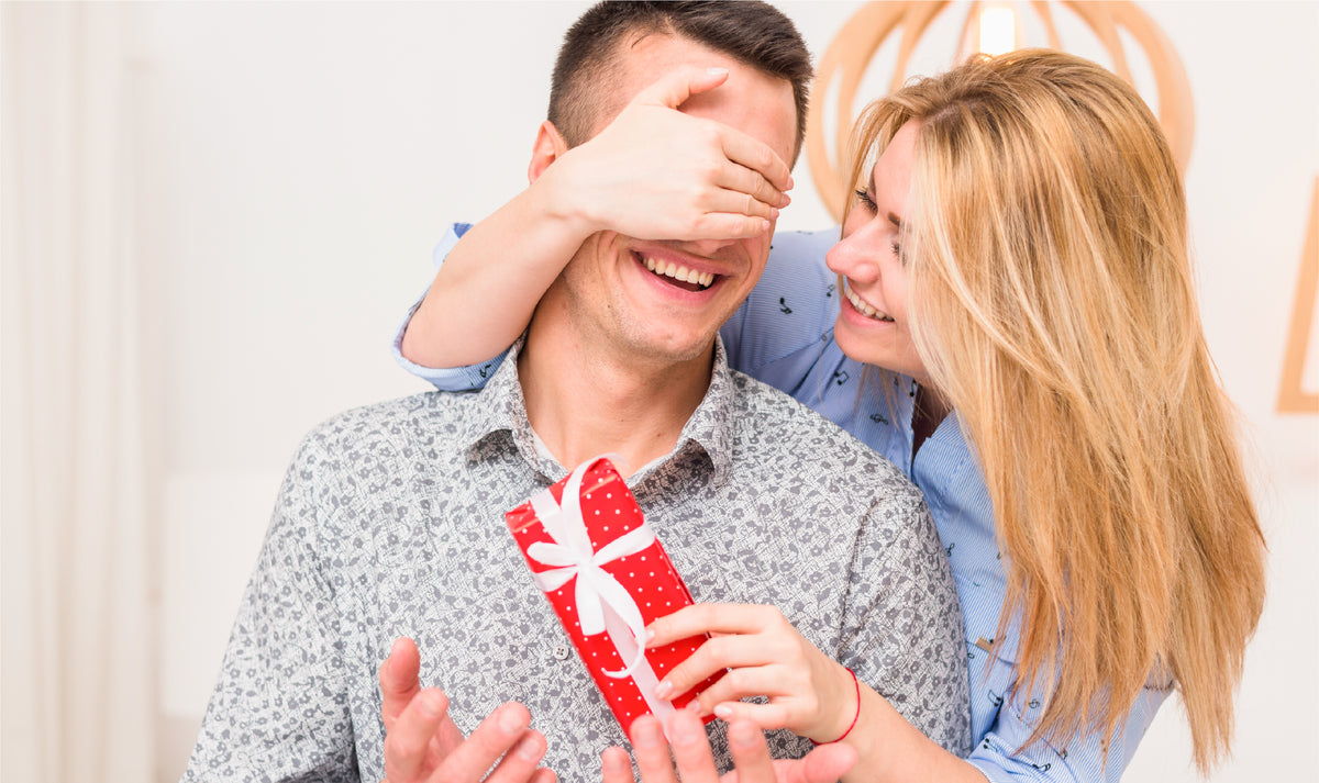 A woman surprises a man by covering his eyes while handing him a red-ribboned gift box, a Robotime Valentine lifestyle shot.