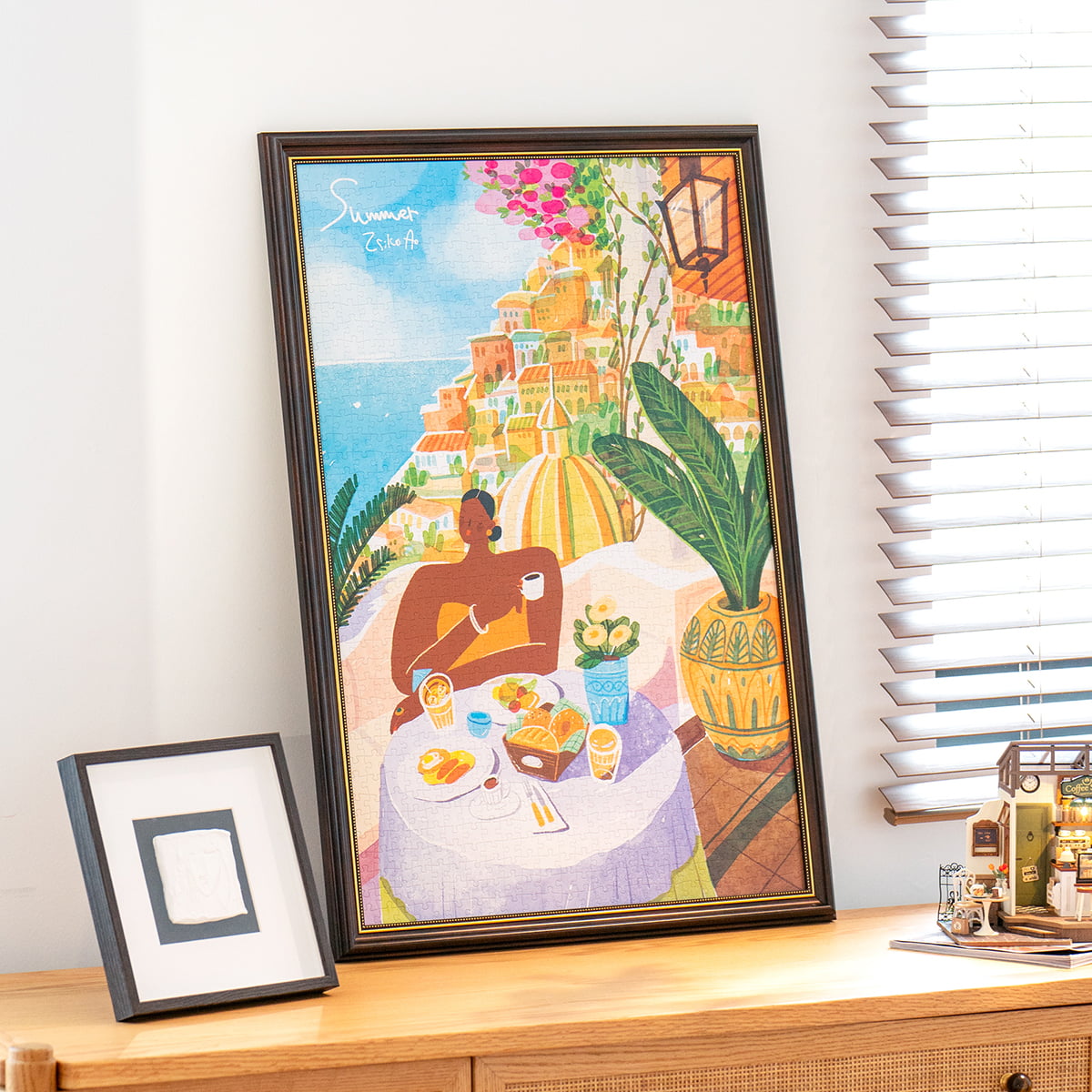 Rolife Amalfi Holiday PT005 completed puzzle in dark frame leaning on wooden shelf beside a smaller photo frame by window