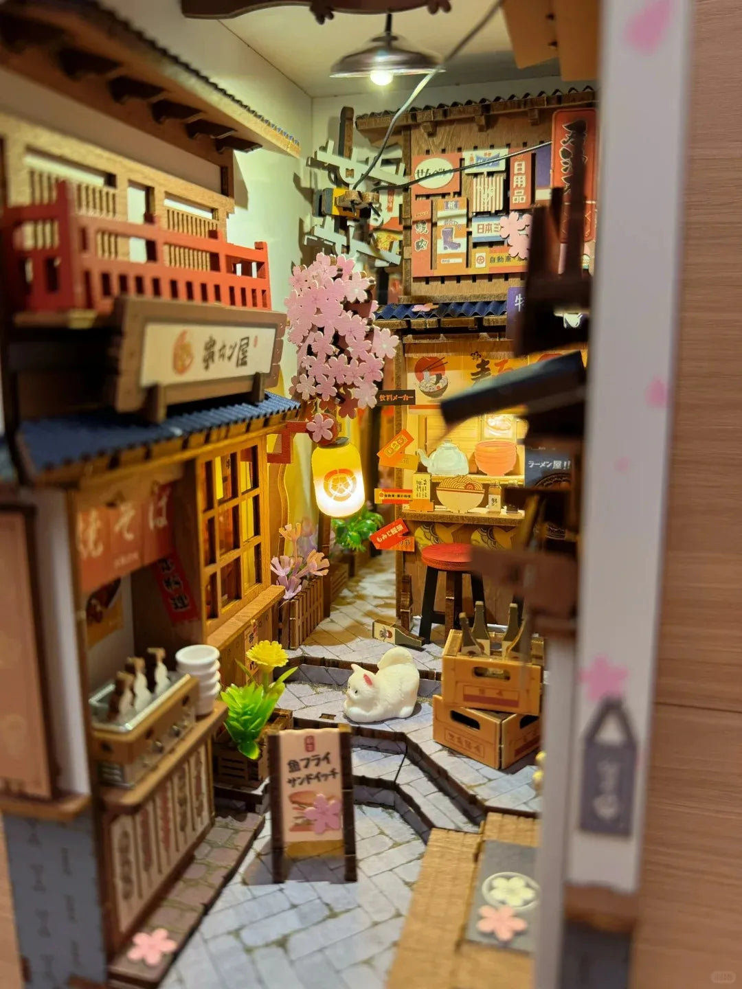 Rolife Sakura Wine Alley TGB11 book nook interior side angle view showing the cherry blossom alley, white cat figurine, and wooden crate with Japanese signage